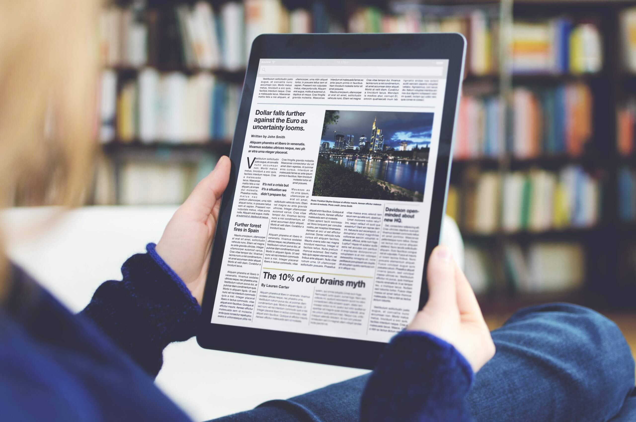 A digital newspaper on a modern tablet computer held by a young woman, who is reading the day’s news. The photo was taken by looking over the model's shoulder. A bookshelf is visible in the background of the picture. The newspaper on the device screen was created by myself in Indesign, I own the copy right of the design and layout. The image of Frankfurt on the device is my own, I own the copy right. The text is all lorem ipsum except for the titles, which are generic and entirely fictional – these were written by myself.